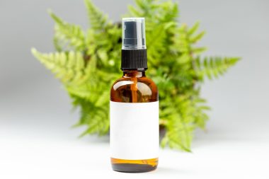 Set of dark amber glass spray bottles with white labels for cosmetic product over green leaves background. Sustainable lifestyle, beauty salon treatment concept, minimalism brand packaging mock up
