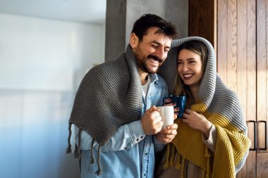 Happy young couple with hot drinks cuddling under warm blanket at home. Weekend people relax love concept.